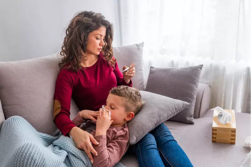 A mother and son on a couch. The mother is looking at a thermometer while the son lays his head on a pillow sitting on her lap while blowing his nose.