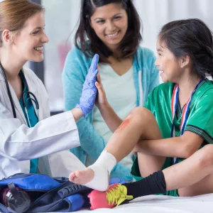 A child being treated for a sprained ankle at the ER, high fiving the ER doctor, while her mom looks on and smiles.