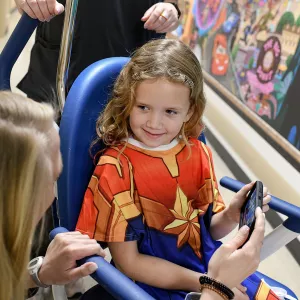 Little girl in a wheelchair at AdventHealth for Children wearing a Captain Marvel gown and using the Disney Team of Heroes mobile app.