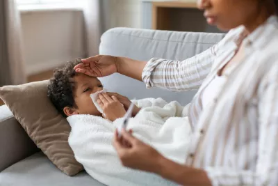 A Small Child Blows His Nose Laying on a Couch While His Mother Checks His Temperature