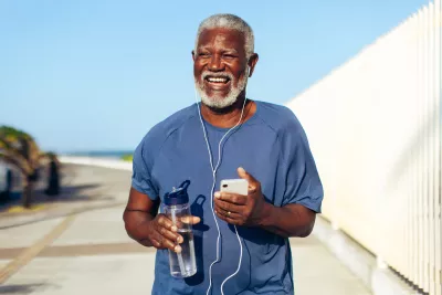 An older black man using headphones plugged into his phone while walking outdoors and holding a water bottle.