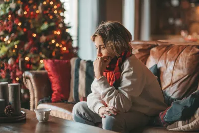 A teen boy sits on a couch at home looking introspectively near a Christmas tree.