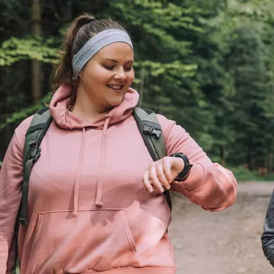 Two Women Go For a Hike Through the Woods as One of Them Checks Their Vitals on Their Smart Watch.