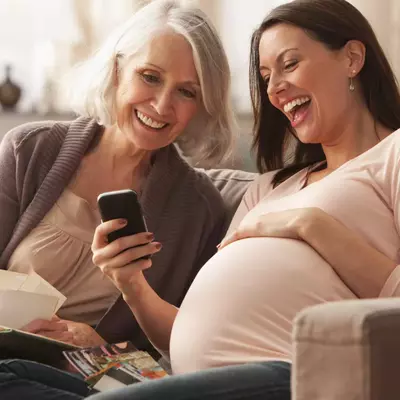 A Mother Sits with Her Pregnant Daughter as they Laugh at Something on a Cell Phone