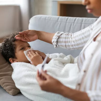 A Small Child Blows His Nose Laying on a Couch While His Mother Checks His Temperature