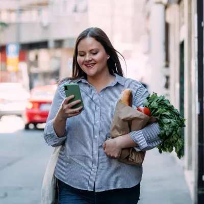 Smiling woman walking and looking at her phone while carrying a bag of groceries.