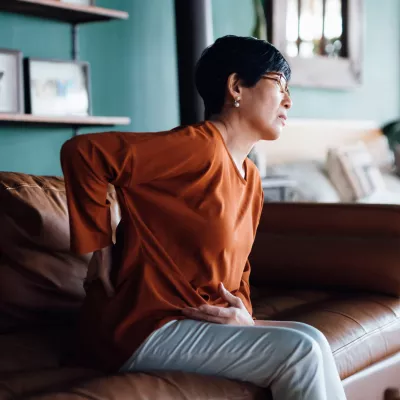 A woman holding her back in pain while sitting on a sofa.