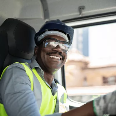 A black man wearing a safety vest, protective glasses, safety helmet and gloves while driving a truck.