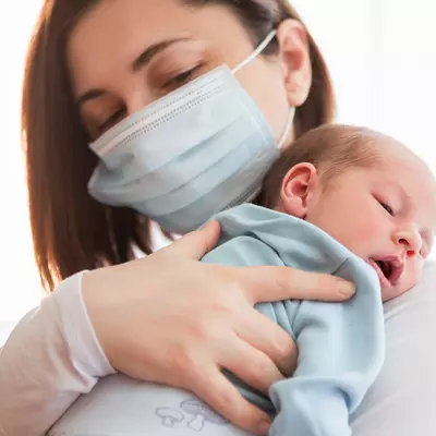 A mom wearing a mask holding her infant.