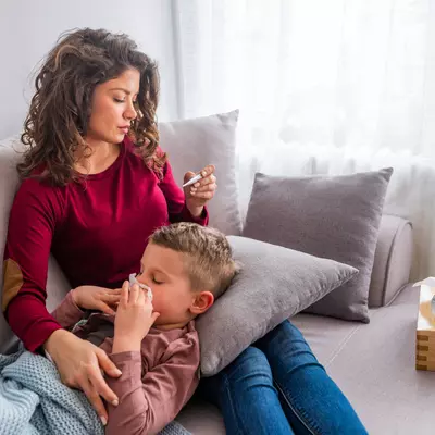 A mother and son on a couch. The mother is looking at a thermometer while the son lays his head on a pillow sitting on her lap while blowing his nose.