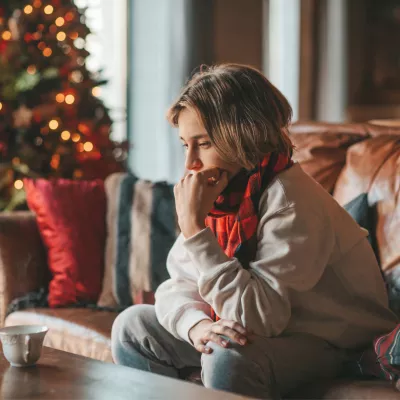 A teen boy sits on a couch at home looking introspectively near a Christmas tree.