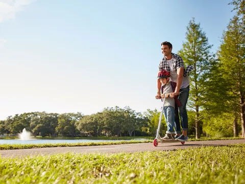 A dad rides a scooter with his son on a sunny day in the park. 