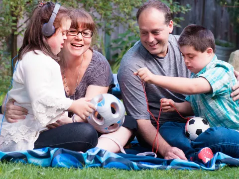 A family of four sitting on a blanket outside, playing with a soccer ball.