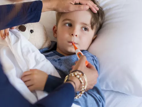 A child in bed with a fever, their parent holding a thermometer in his mouth.