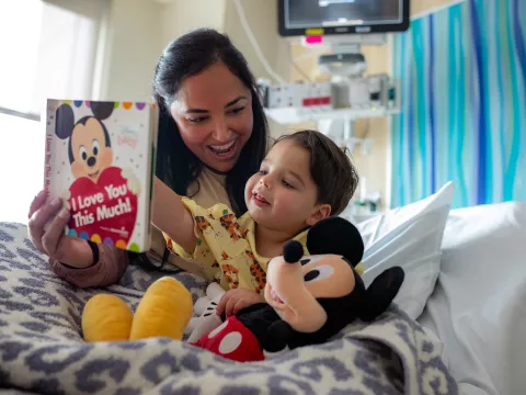 An AdventHealth for Children patient and his mother read a Mickey Mouse book together in a hospital bed with a Mickey plush toy.