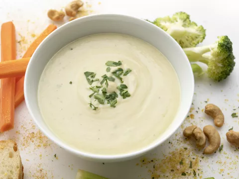 White bowl of cream soup next to carrot sticks, broccoli, and cashews