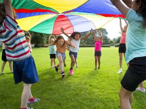 A group of children outside playing with a large multicolored parachute.