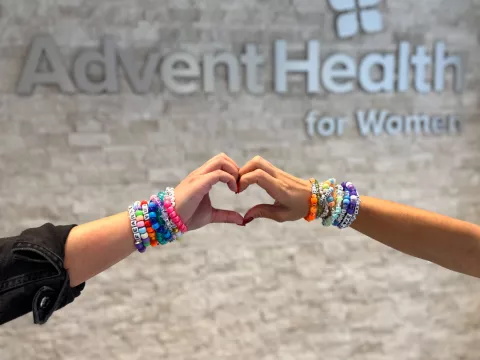 Two hands with friendship bracelets on the wrists form a heart in front of the AdventHealth for Women sign.