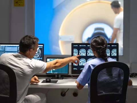 A male and female imaging technician looking at CT scans of a patient's head in the foreground while another male technician is prepping the patient in the CT machine in the background.