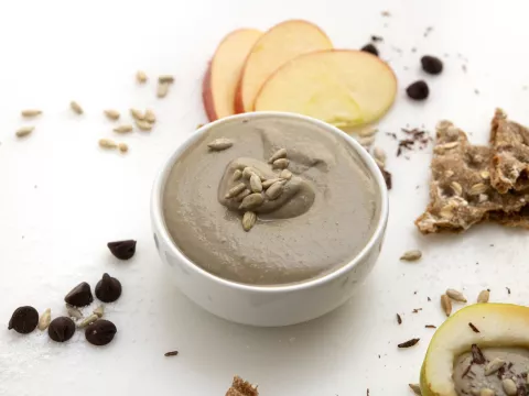 White bowl of brown sunflower seed butter next to apples and crackers.