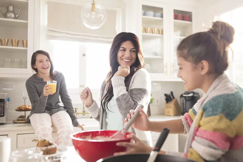 A Caucasian mom dances around the kitchen with her two daughters.