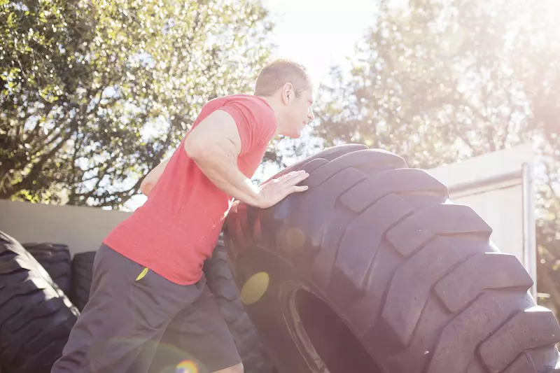 A young Caucasian man exercises outdoors by flipping large tires.