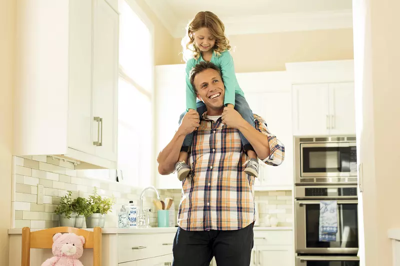 A Caucasian father carries his young daughter on his shoulders in their home.