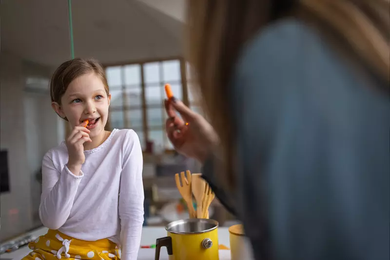 A Mother Hands Her Daughter Carrot Sticks to Eat in the Kitchen.