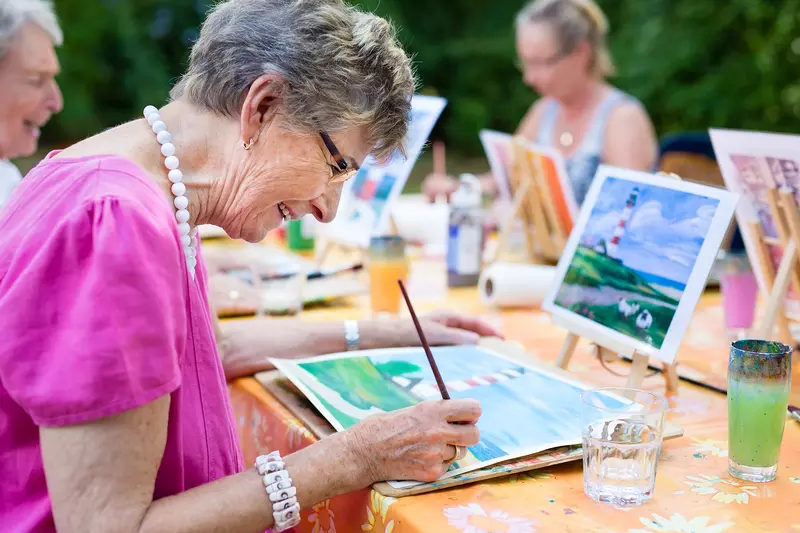 A Grandmother Paints a Watercolor Painting Based on an Example on a Tablet.