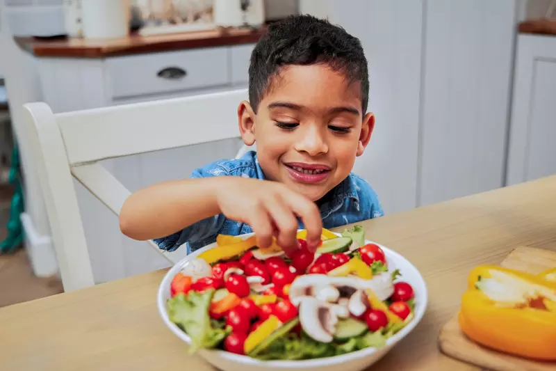 A Young Boy Grabs Some Vegetables From a Salad on a Kitchen Table.