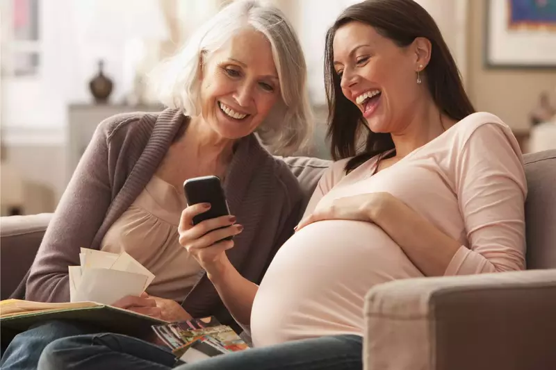 A Mother Sits with Her Pregnant Daughter as they Laugh at Something on a Cell Phone