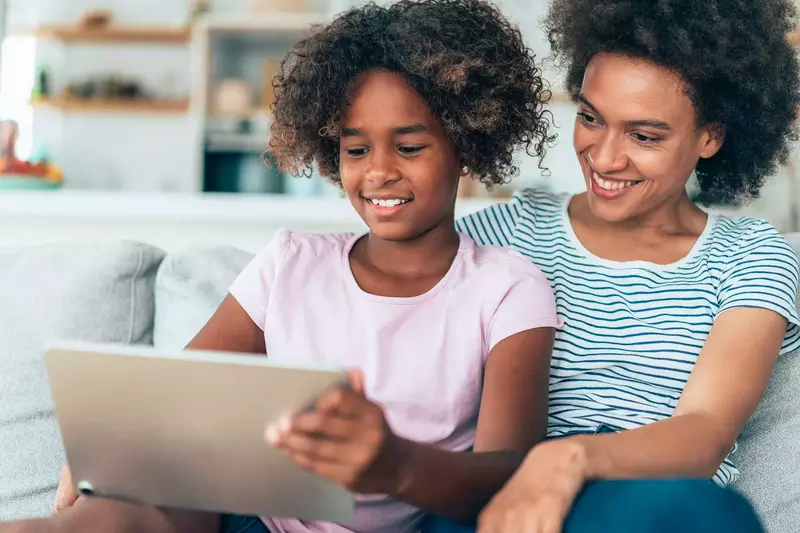 A MOther and Daughter Smile as They Sit Together and Surf the Internet on a Tablet.
