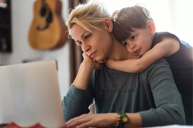 A Mom Stares at A Computer Screen While Her Young Son Hugs Her From Behind