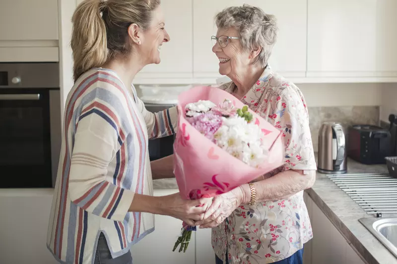 Middle aged woman giving an elderly woman light colored flowers in pink wrapping. They are both standing in a white kitchen.