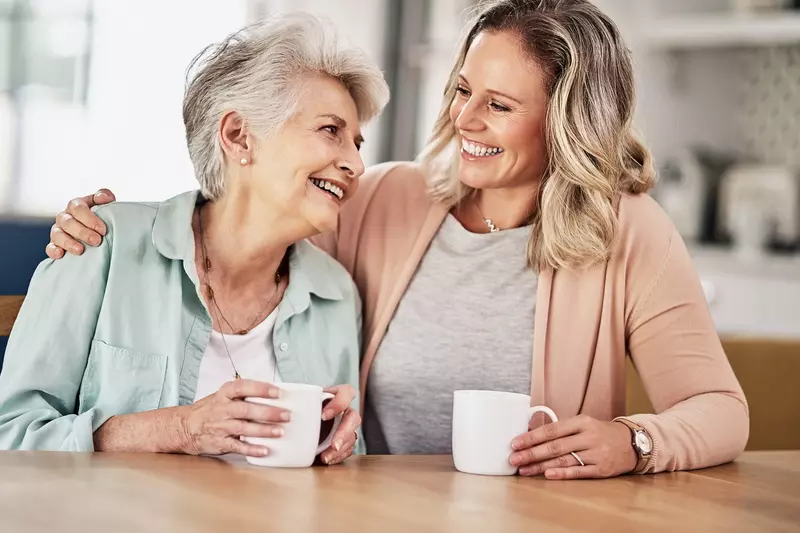 Adult woman with her arm around her mother, both smiling and holding white mugs.