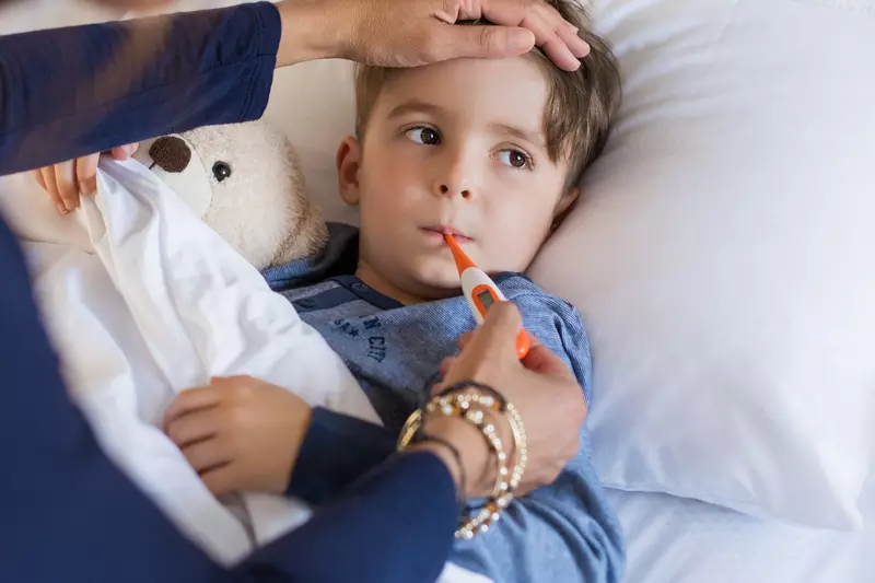 A child in bed with a fever, their parent holding a thermometer in his mouth.