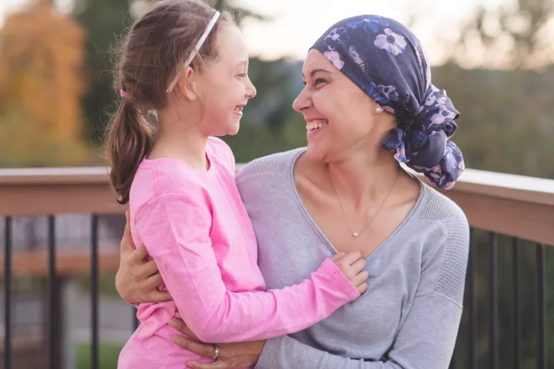 A mother and cancer survivor is sitting, with one arm around her daughter.