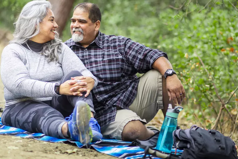 A senior couple is sitting on the ground outdoors enjoying a picnic after a hike.