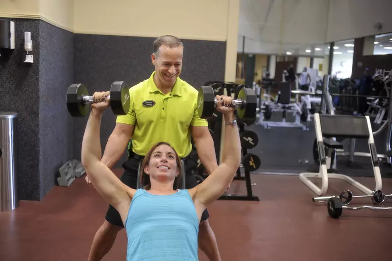 A personal trainer stands behind woman a woman who is laid out on a bench, bench pressing dumbbells.