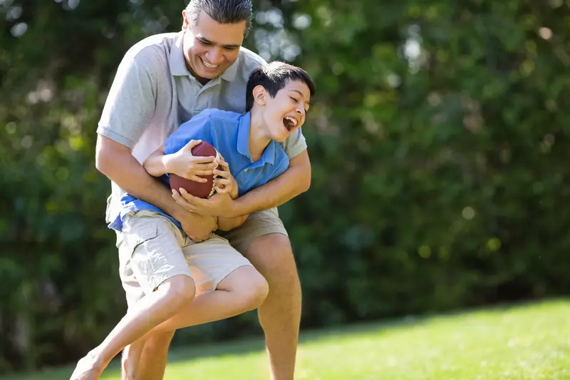 A father playfully tackles his son as they play football outside.