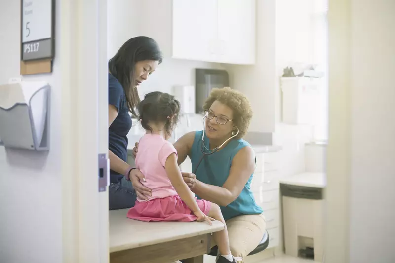 A female AHS doctor listens to a little girl's heart in the exam room while her mother comforts her