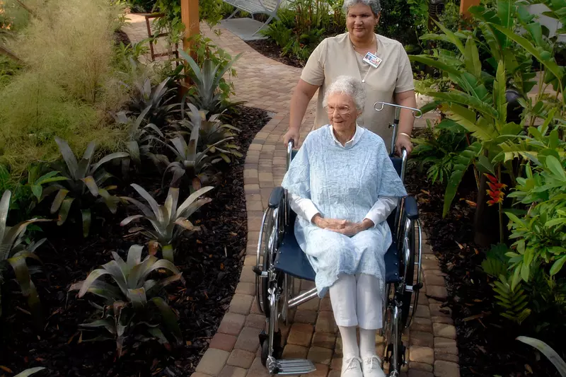 Elder woman in a wheelchair being pushed down a path through a garden.