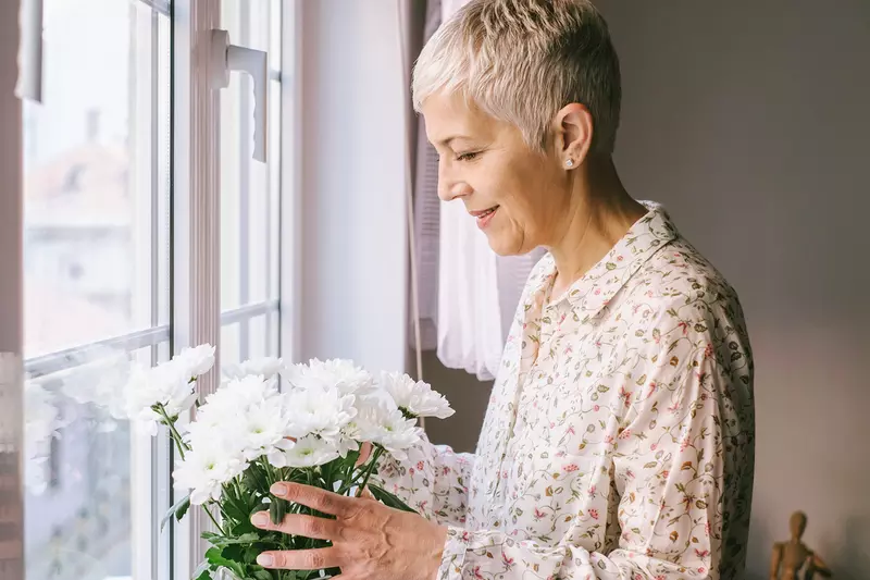 An adult caucasian woman arranges a vase of flowers by the window.