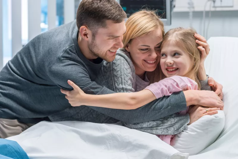 A family hugging their young daughter in her hospital bed.