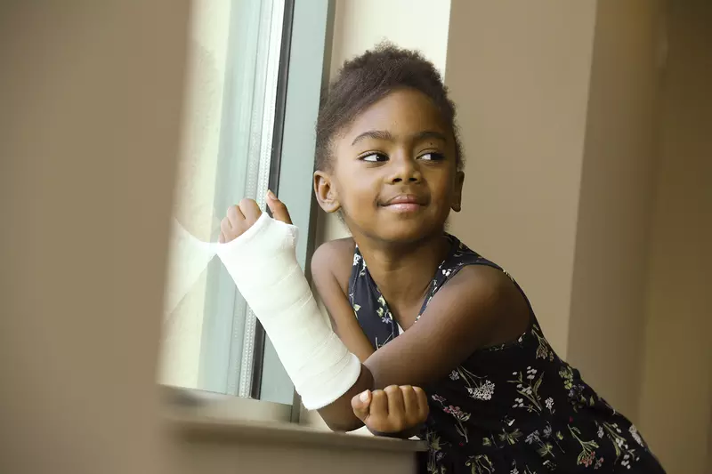 A little girl in a cast smiles while sitting next to a windowsill