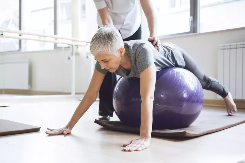 A Caucasian woman balances herself on an exercise ball.