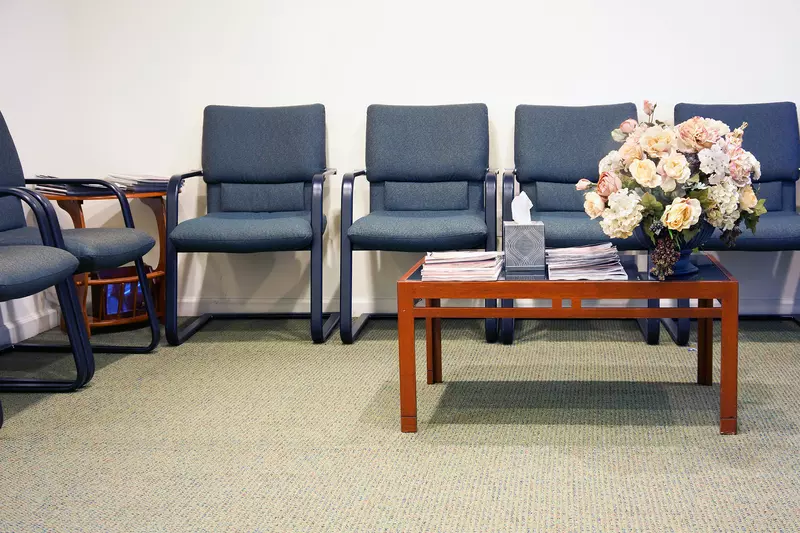 Hospital waiting room with blue chairs and a brown coffee table displaying flowers.