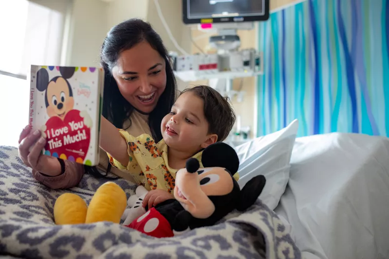 An AdventHealth for Children patient and his mother read a Mickey Mouse book together in a hospital bed with a Mickey plush toy.