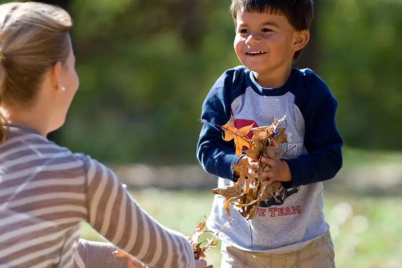 A boy plays happily in the fallen leaves with his mother.
