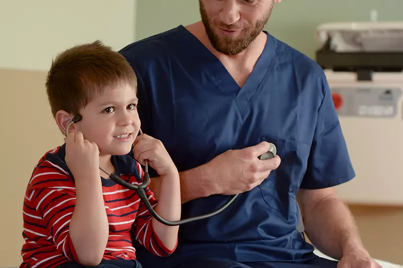 A male nurse lets his child patient listen to a stethoscope.